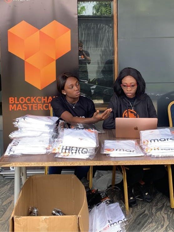 Timonwa is sitting behind a table alongside another female volunteer. She is working on a laptop. The Mara Blockchain Masterclass banner is behind them.